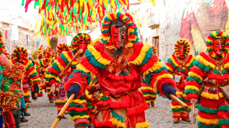 Gros plan sur des Caretos de Podence portant des masques de métal au nez pointu et des costumes à franges colorées courant dans une rue pavée.