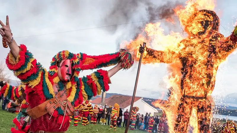 Gros plan sur des Caretos de Podence portant des masques de métal au nez pointu et des costumes à franges colorées sautant devant une statut en feu