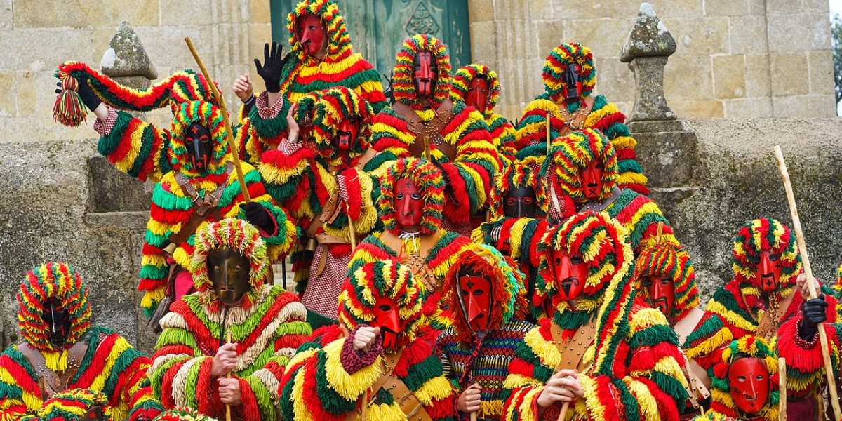 Gros plan sur des Caretos de Podence portant des masques de métal au nez pointu et des costumes à franges colorées sautant dans une rue pavée.