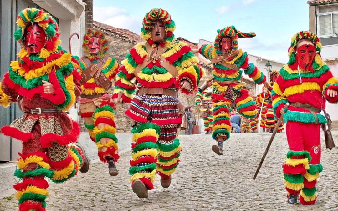 Groupe de Caretos masqués portant des costumes à franges colorées et des cloches, en train de courir et de sauter dans une ruelle pavée typique du village de Podence.