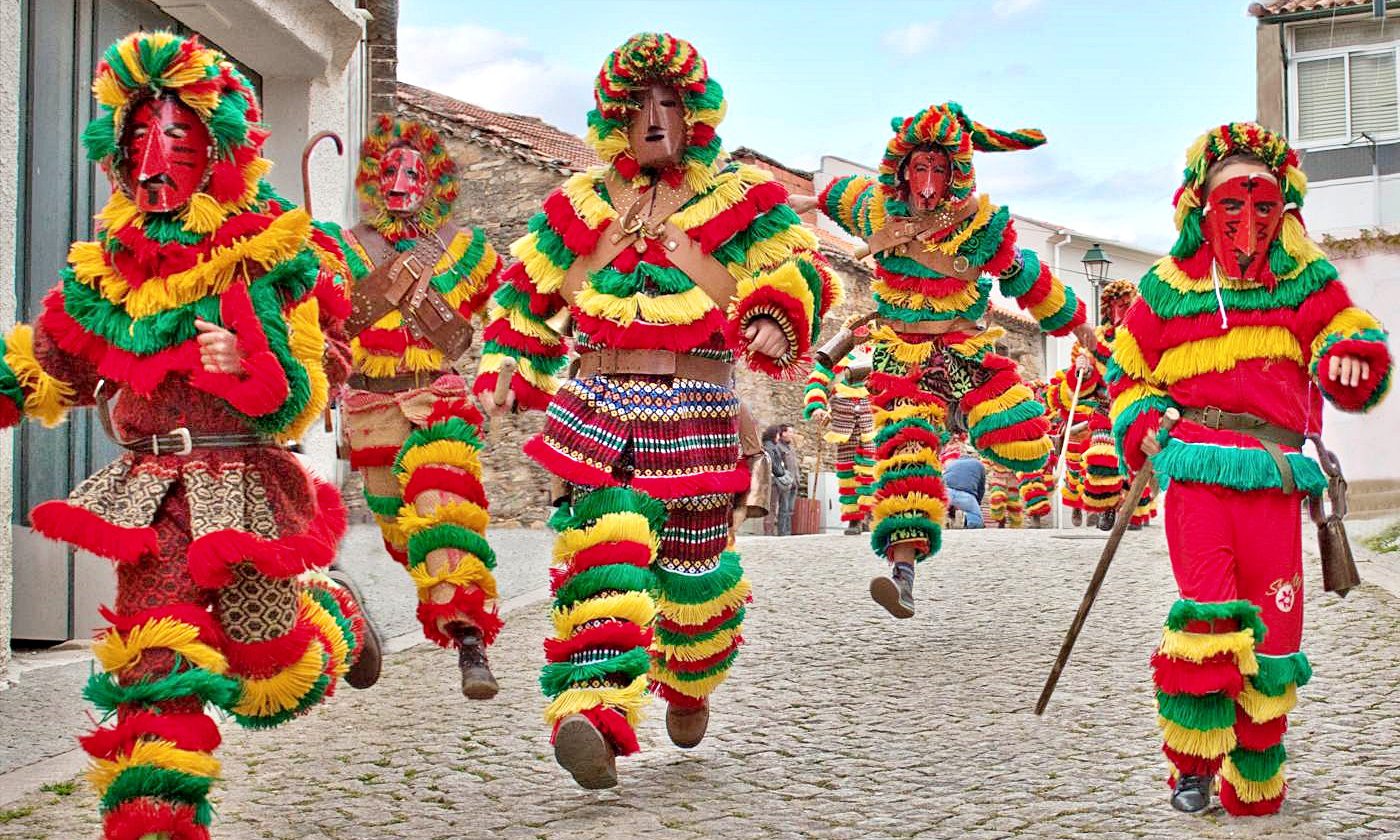 Groupe de Caretos masqués portant des costumes à franges colorées et des cloches, en train de courir et de sauter dans une ruelle pavée typique du village de Podence.